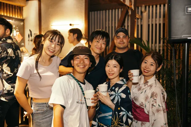 A group of smiling young people holding drinks and posing together at a warm evening venue