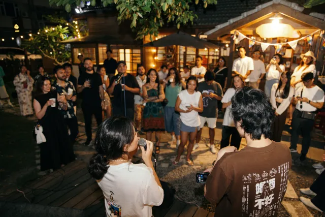 A crowd of young attendees gathers outdoors at night around a speaker under warm string lights