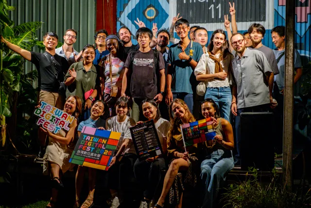 A diverse group of around twenty people pose cheerfully outdoors at night holding Taiwan Digital Fest 2025 booklets