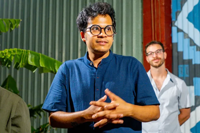A speaker in a blue linen shirt gestures with clasped hands while addressing an outdoor evening event