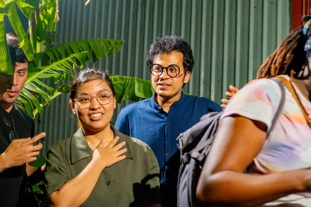 Four people conversing outdoors at night near tropical foliage, smiling warmly