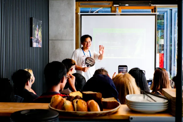A presenter waves to a seated audience in a cafe with bread on the table and a projection screen behind him