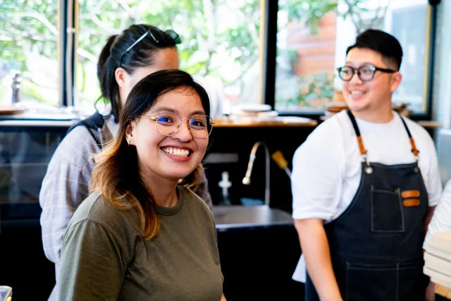 A smiling woman with glasses turns toward the camera inside a cafe kitchen, with two apron-wearing staff behind her