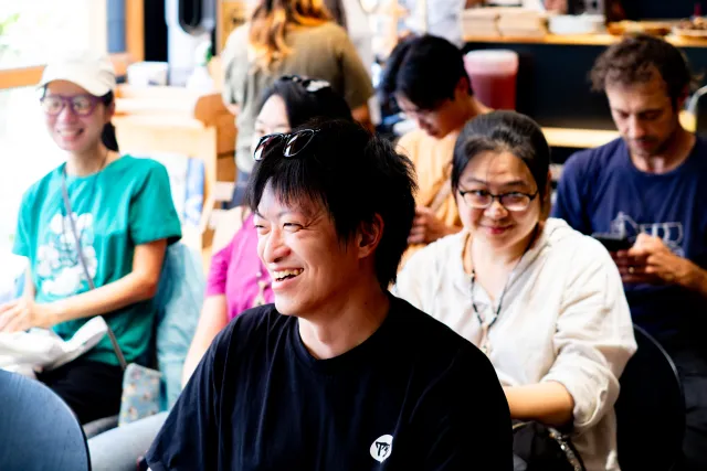 A smiling man seated among a group of people laughing together in a casual indoor gathering