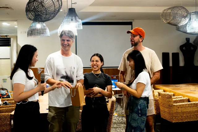 Five people laughing and exchanging a gift bag at an indoor event