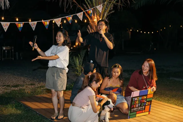 Emcee and host perform on a wooden stage at an outdoor night market, while seated guests laugh nearby