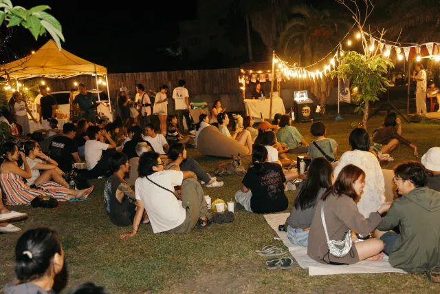 Crowd of young people sitting on grass at an outdoor night market under string lights