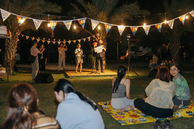 Audience sits on picnic blankets at night watching presenters on a small outdoor stage strung with festive lights