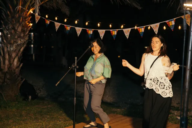 Two women dancing joyfully outdoors at night under string lights and colorful pennant flags