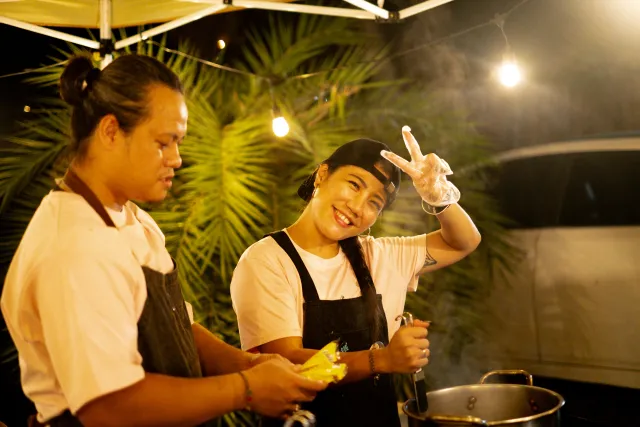 Two vendors in aprons smile and pose while cooking at an outdoor night market stall