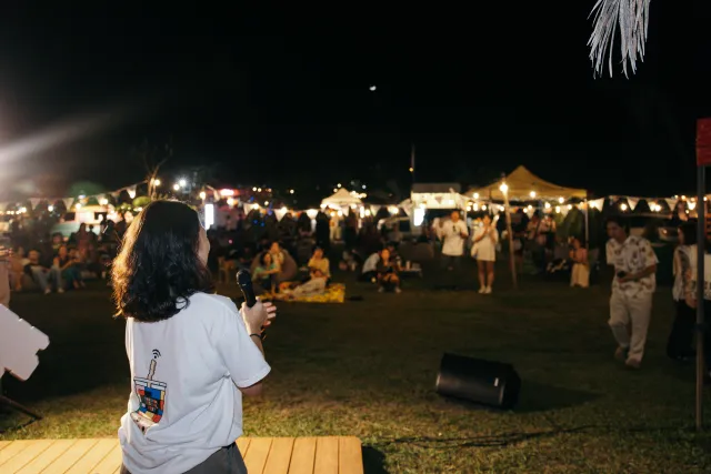 A host holding a microphone addresses a lively outdoor night market crowd under string lights