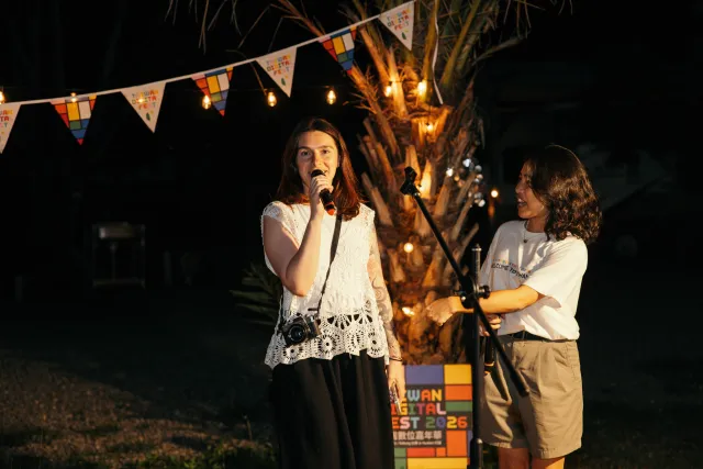 A woman speaks into a microphone outdoors at night under string lights and colorful bunting