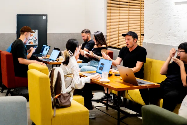 A group of people collaborating on laptops at cafe tables, one man reacting with surprise