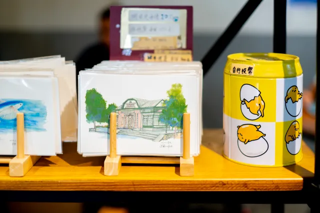 Illustrated postcards of a historic building displayed on a wooden table beside a donation tin