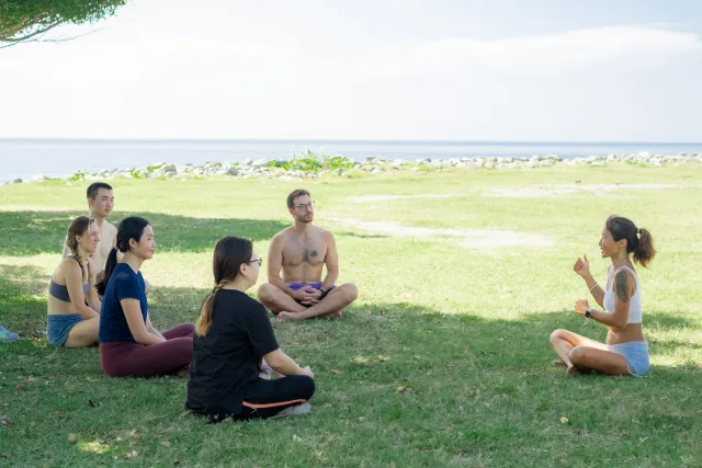 A small group sits cross-legged on a grassy waterfront lawn, listening to an instructor during a morning yoga session