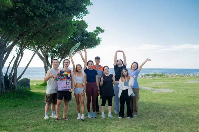 Group of nine people posing cheerfully on a grassy seaside lawn under trees