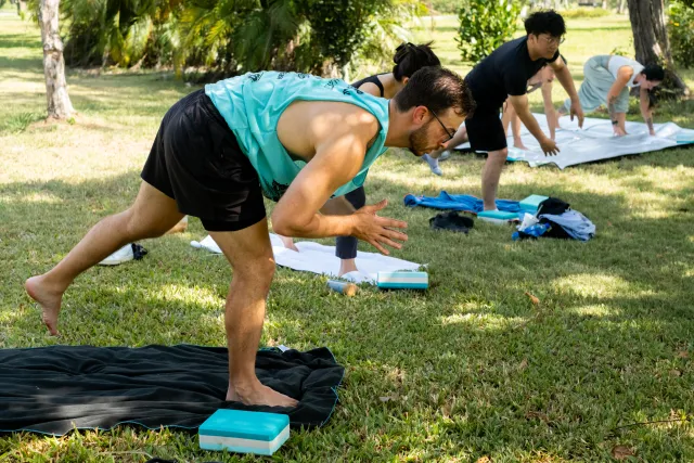 participants hold a forward fold yoga pose on grass mats outdoors under trees