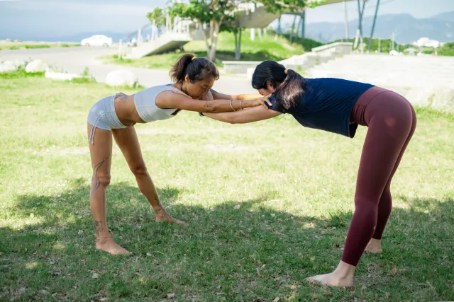 Two women face each other bowing forward and clasping wrists in a partner stretch on a grassy lawn