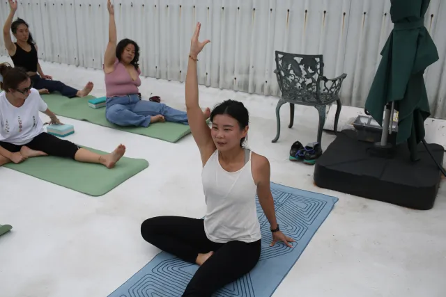 Women seated on yoga mats raise one arm in a stretch during an outdoor morning session