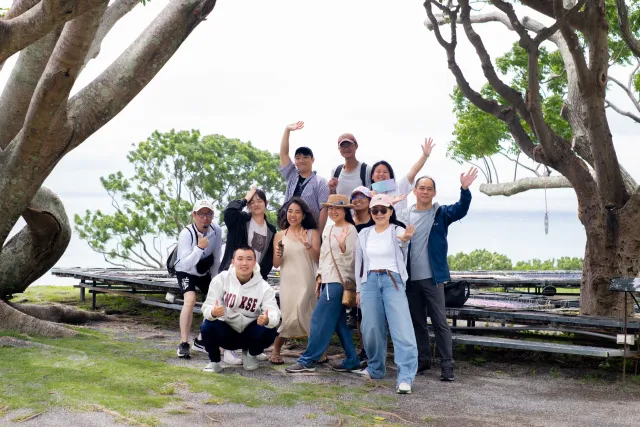 A group of about ten people pose and wave cheerfully under large trees by the ocean