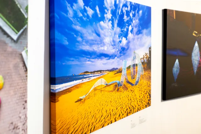 framed beach photo on gallery wall showing colorful art installations on golden sand under blue sky