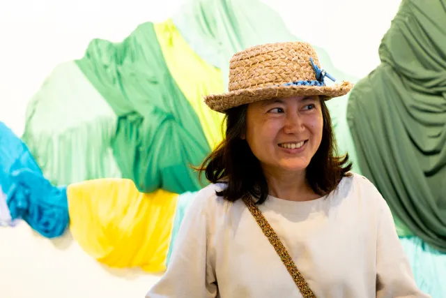 A smiling woman in a straw hat poses in front of a colorful draped fabric art installation