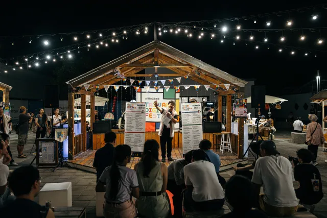 A speaker addresses a seated crowd at an outdoor night market opening stage under string lights