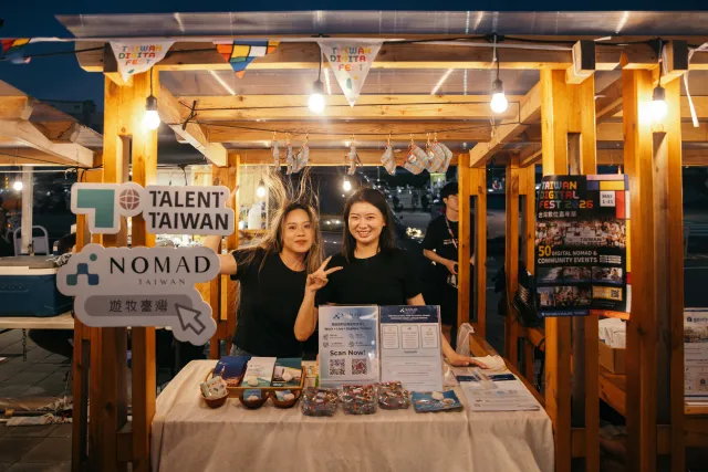Two women smile and pose at a Talent Taiwan / Nomad Taiwan booth during an outdoor night market event