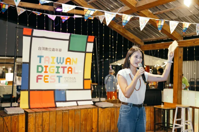 A woman speaks into a microphone on stage before a Taiwan Digital Fest backdrop at an evening outdoor event