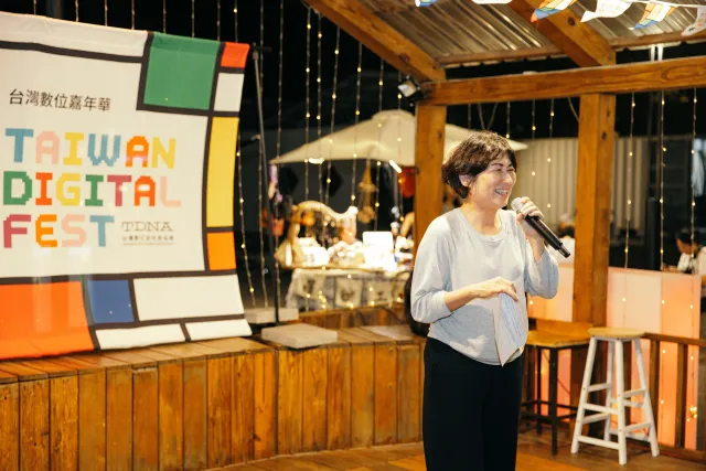 A woman speaks into a microphone at an indoor opening event beside a Taiwan Digital Fest banner