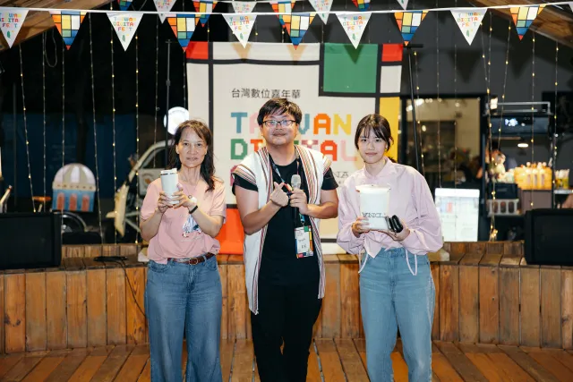 Three people pose on a wooden stage in front of a Taiwan Digital Fest backdrop, the center person holding a microphone