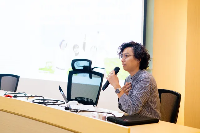 A speaker holds a microphone and presents at a conference table with a projected slide behind them