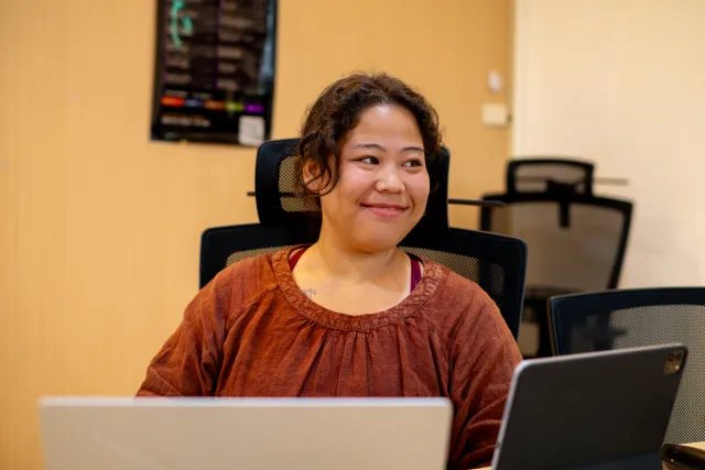 A woman sits at a laptop in a seminar room, smiling and looking off to one side