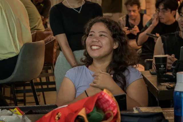 A young woman smiles warmly at an indoor evening event, seated among other attendees