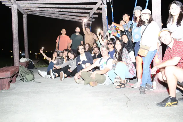 A large group of people pose together outdoors under a wooden pergola at night
