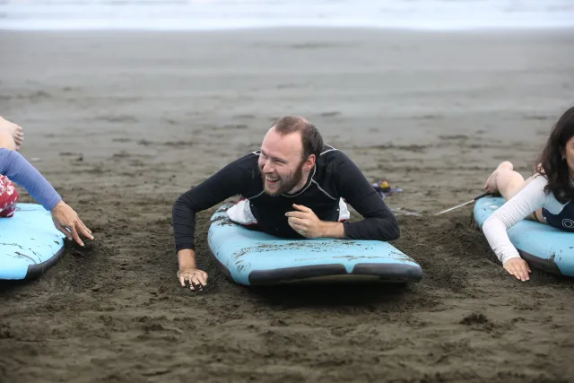 A man in a wetsuit lies prone on a surfboard on a sandy beach, smiling and chatting with others beside him