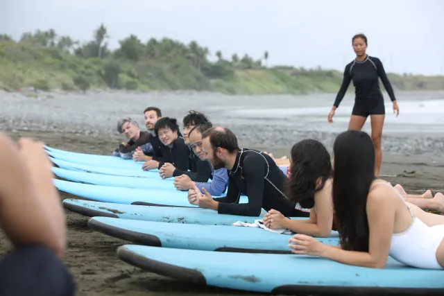 Surf students lie prone on blue boards on a dark sand beach while an instructor stands nearby