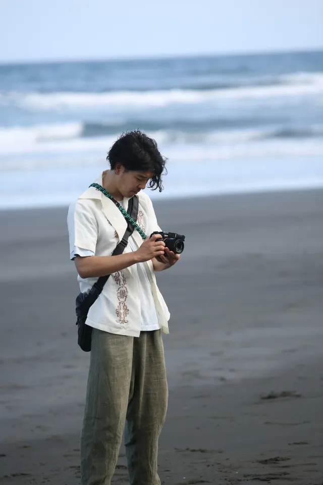 A person reviews photos on a camera while standing on a dark sand beach with waves behind
