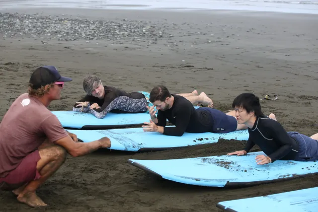 an instructor crouches on the beach demonstrating surfboard technique to three prone students