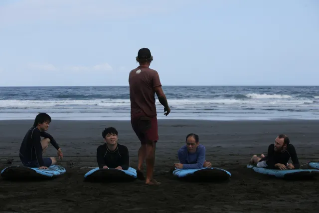 Surf instructor standing on black sand beach briefing four students lying on surfboards