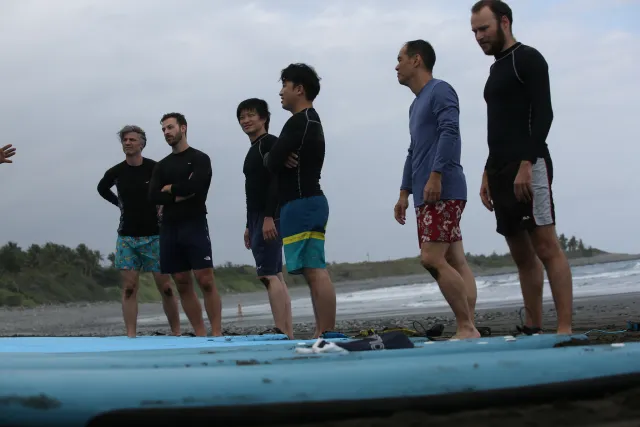 Six participants in rash guards stand on a dark sand beach listening before a surf lesson
