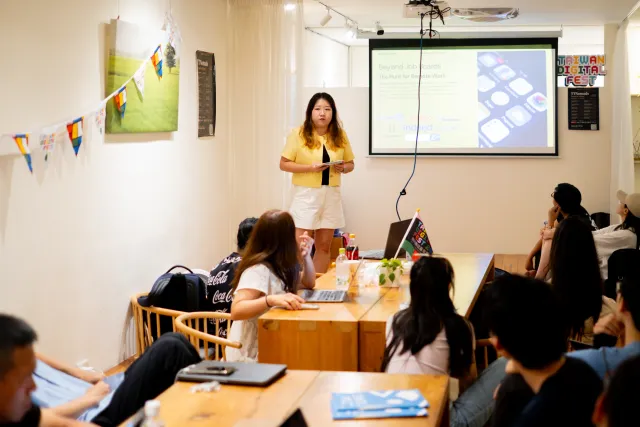 A woman in a yellow top presents slides to a seated audience in a cozy indoor venue