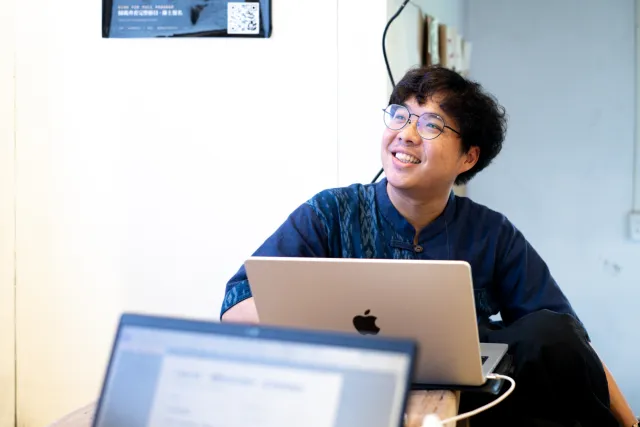A smiling man with glasses sits behind a laptop, looking up cheerfully during a talk session