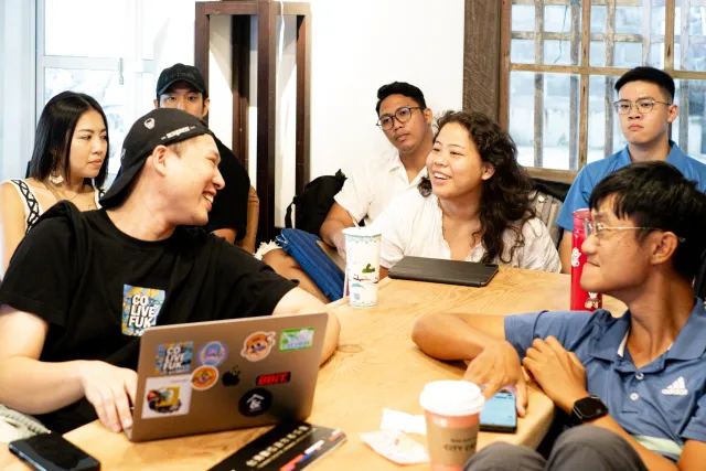 A group of young people laugh and chat together around a wooden table during a workshop session