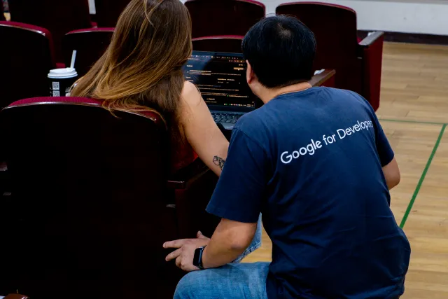 Two attendees huddle over a laptop in an auditorium, one wearing a Google for Developers shirt