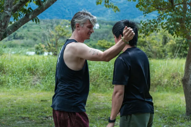 Two men face each other outdoors as one places a hand on the other's head during a workshop activity