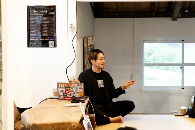 A speaker sits cross-legged on a platform, gesturing while presenting to an indoor workshop audience
