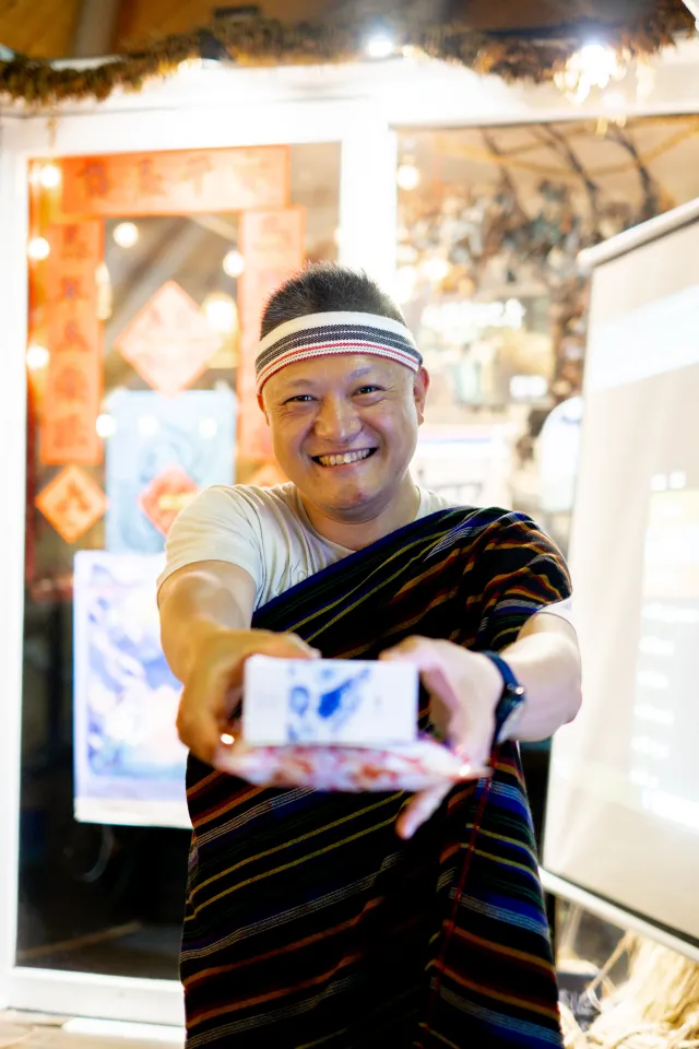 smiling man in traditional striped cloth and headband holds a small printed photo toward the camera