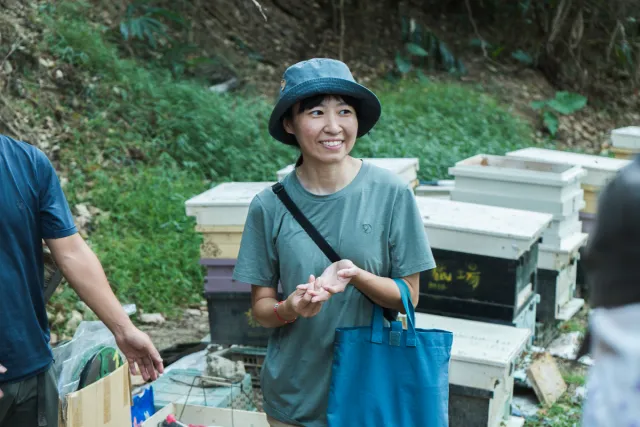 A smiling woman in a bucket hat stands among rows of beehives outdoors