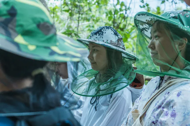Participants wearing insect-net hats listen attentively outdoors among trees